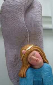 wooden sculpture of a female figure wearing a circlet on her head and in a blue outfit. She seems somewhat slumped