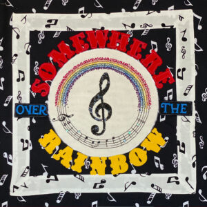 stitched fabric square with borders showing musical symbols. In the centre is a treble clef in a circle with a rainbow above, and surrounded by the works somewhere over the rainbow spelled out in large letters. Part of the Covid Chronicle