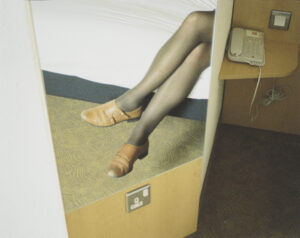 a photography focusing on a pair of legs crossed, wearing sheer black tights and brown shoes. The sitter's body is unseen, they are sitting down behind something.