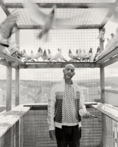 Black-and-white photograph by Craig Easton, a lead image for his exhibition "Is Anybody Listening?". Featuring a bald man standing in a large pigeon coop filled with birds.