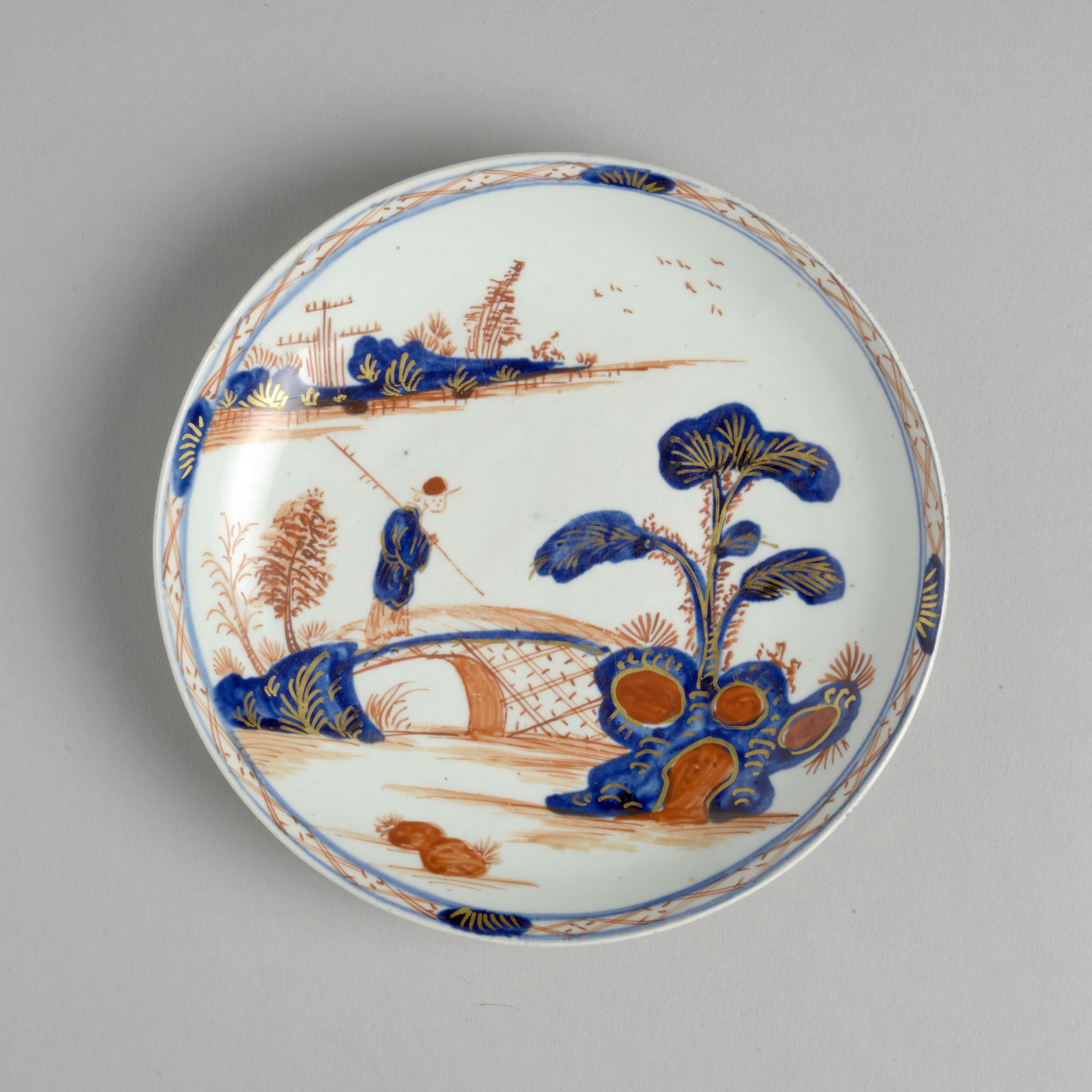 A white saucer decorated with a design which is clearly inspired by willow pattern - a man crossing a chinese bridge - but in orange as well as blue.