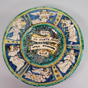 a della robbia plate painted in green, blue and yellow. Around the outside are angels and in he middle is reads peace on earth, good will towards men