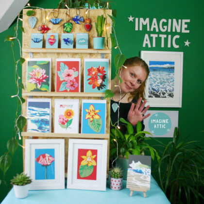 photograph of a market stall, with the words "imagine attic" written on the wall behind. A display stand shows a range of prints and cards, and a woman is popping her head out from behind and waving.