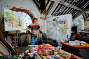 Photograph of the artist Rob Davies (an adult male with dark hair and glasses, wearing a beige jumper with a red and green stripe) in his studio, in front of a window. He is standing behind a table covered in various art supplies and between two of his works, both landscape watercolours