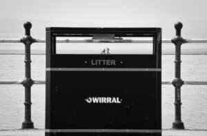 black and white photograph taken on West Kirby waterfront overlooking the Marine Lake, part of Jonathan Cooper's exhibition series Life Line. Photo shows a bin and we can see right through the gap for rubbish to the other side of the lake, where we see two figures, one pushing another in a wheelchair. The effect is of tiny people walking on the edge of the bin.