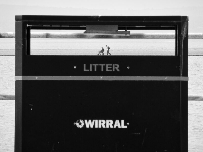 black and white photograph taken on West Kirby waterfront overlooking the Marine Lake, part of Jonathan Cooper's exhibition series Life Line. Photo shows a bin and we can see right through the gap for rubbish to the other side of the lake, where we see two figures, one pushing another in a wheelchair. The effect is of tiny people walking on the edge of the bin.