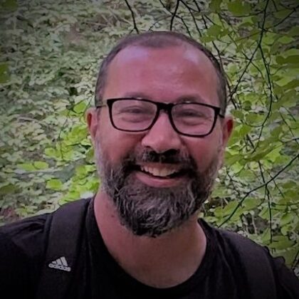 photograph of a white man with short hair, a short beard and black glasses, who is smiling. He is standing in front of a large bush which fills the entire background