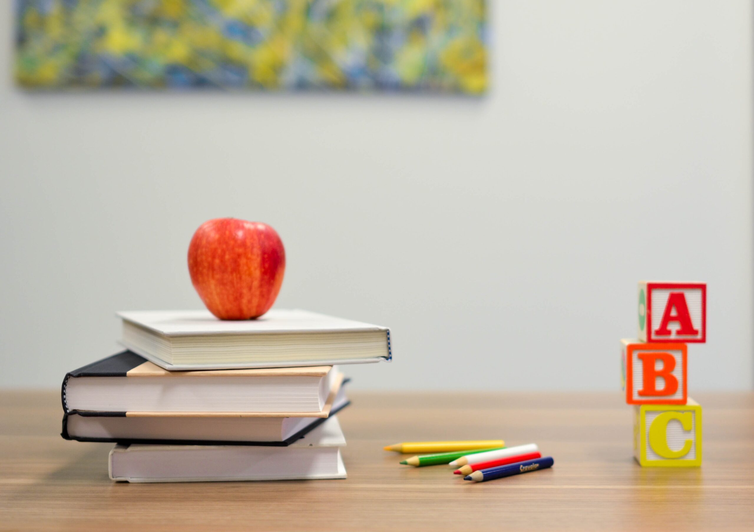 an apple sitting on a pile of 4 books on a desk, there's also some coloured pencils on the tables and stacked bricks reading ABC