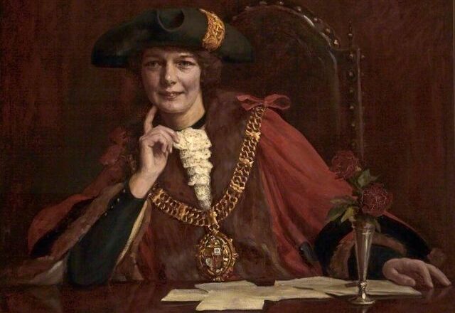 a portrait of a woman wearing full mayoral regalia - red cloak, chain and tricorner hat, sitting at a desk