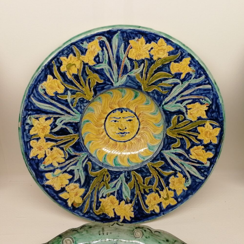 a decorative plate. In the centre is a bright yellow sun with a human face. Around the outside of this is a dark blue background decorated with yellow and green daffodils